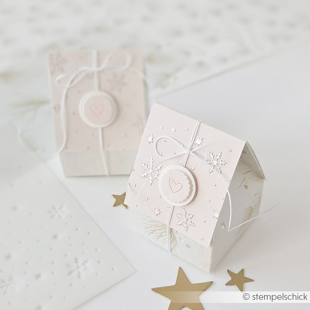 Zwei hellrosa, hausförmige Geschenkboxen, verziert mit Schablonenmotiven Rieselnde Schneeflocken und Herzen.