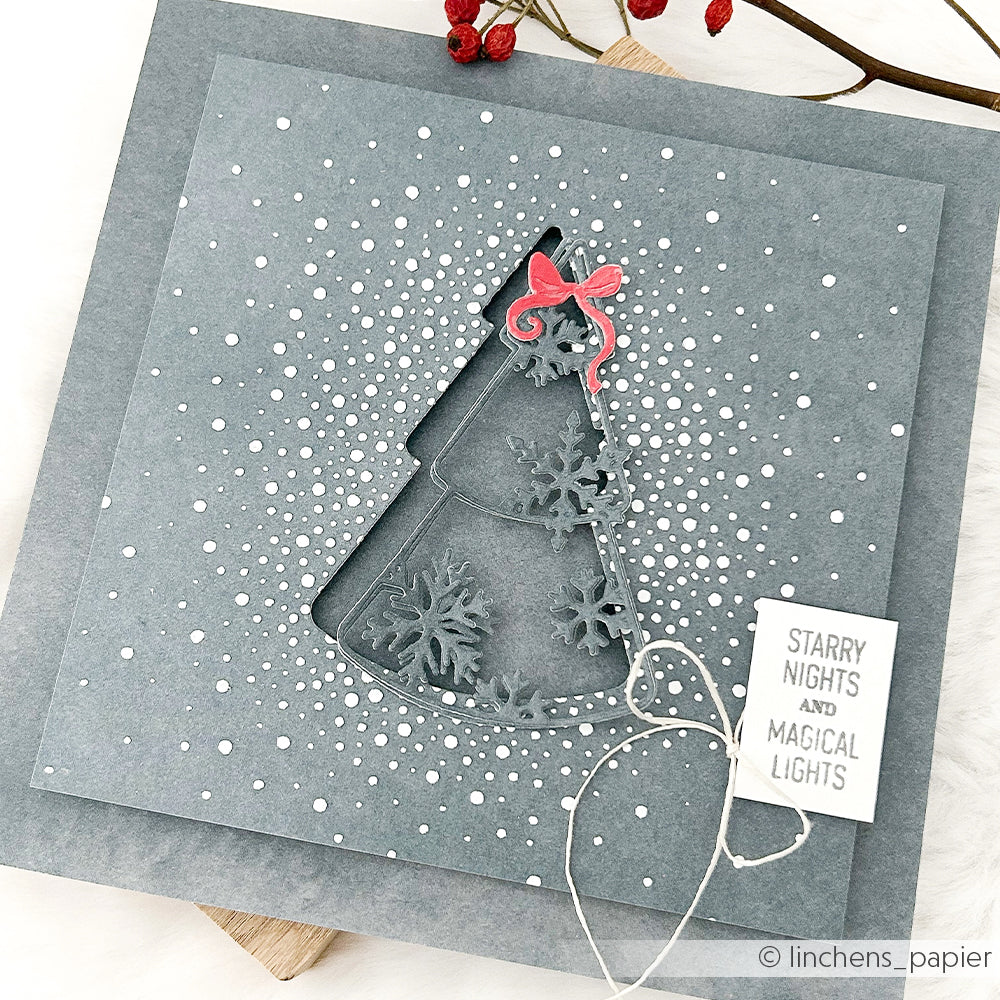 Graue Weihnachtskarte mit einem ausgestanzten Stencil Tannenbaum-Motiv, Schneeflocken, einer roten Schleife und einem Starry Nights-Anhänger.