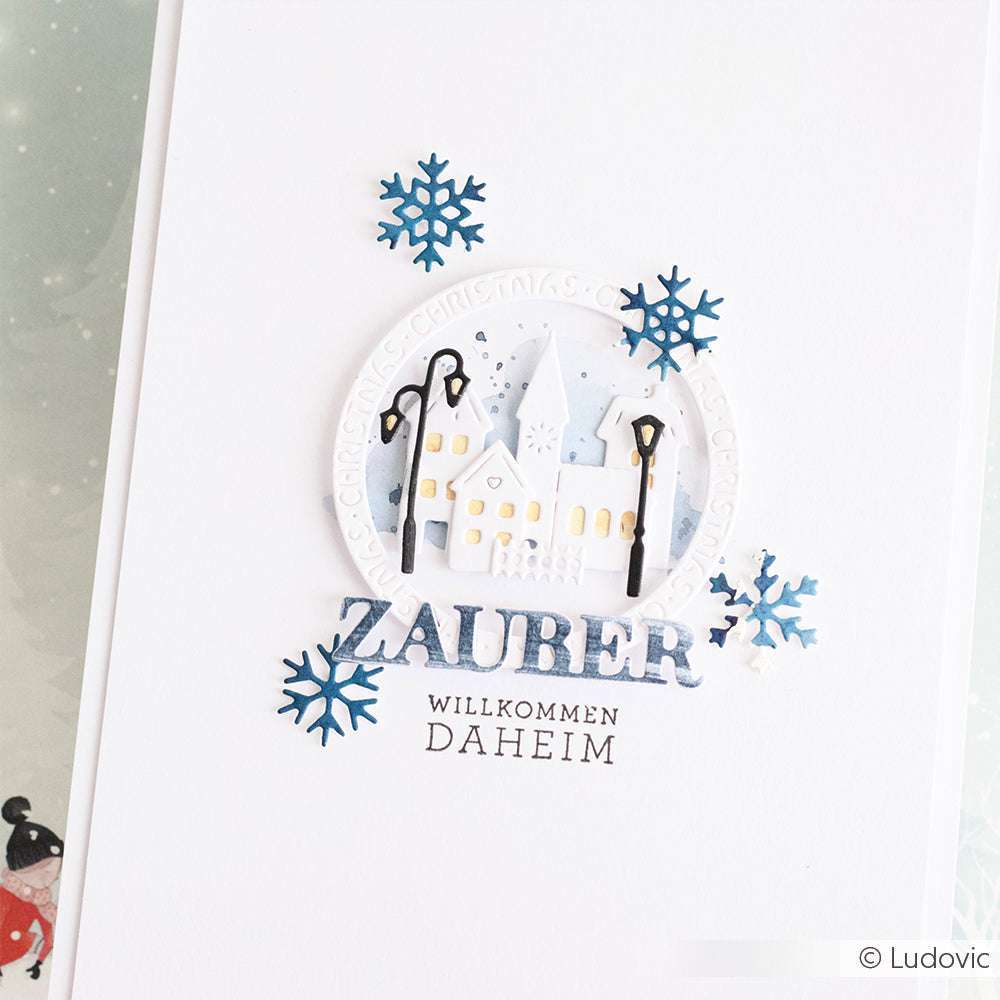 Die handgefertigte Winterkarte von Alexandra Renke zeigt ein verschneites Dorf, blaue Schneeflocken und den deutschen Text "Zauber Willkommen Daheim", gestempelt mit einem hochwertigen Photopolymer-Stempel für scharfe Details.