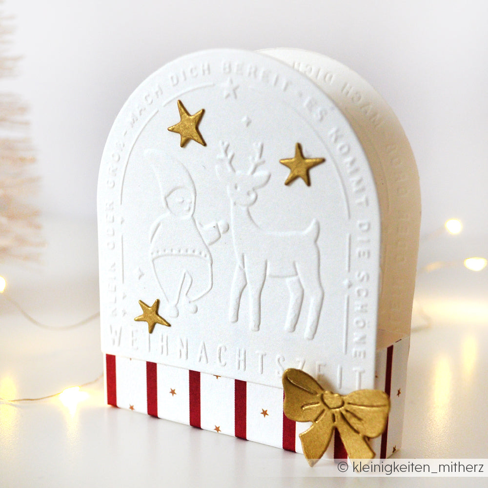 Weiße geprägte Weihnachtskarte mit dem Schneide- und Prägefolder von Alexandra Renke, mit goldenen Sternen, einer goldenen Schleife, Rentieren und einem Wichtel aus Wichtelgrüße sowie roten Streifen für einen festlichen Touch.