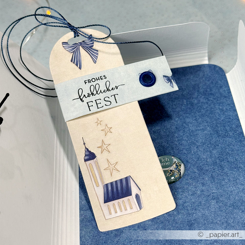Alexandra Renke Geschenkanhänger mit einer Kirche und Sterne Illustration mit "Frohes Fest", gebunden mit blauer Schnur, vor einem blauen Hintergrund - perfekt für die Weihnachtszeit.
