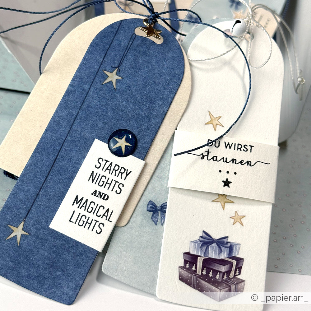 Blaue und weiße handgefertigte Geschenkanhänger mit Sternen, festlichem Text und einem Stapel Geschenke, die mit Alexandra Renke-Klarsichtstempeln für gestochen scharfe, detaillierte Designs hergestellt wurden.