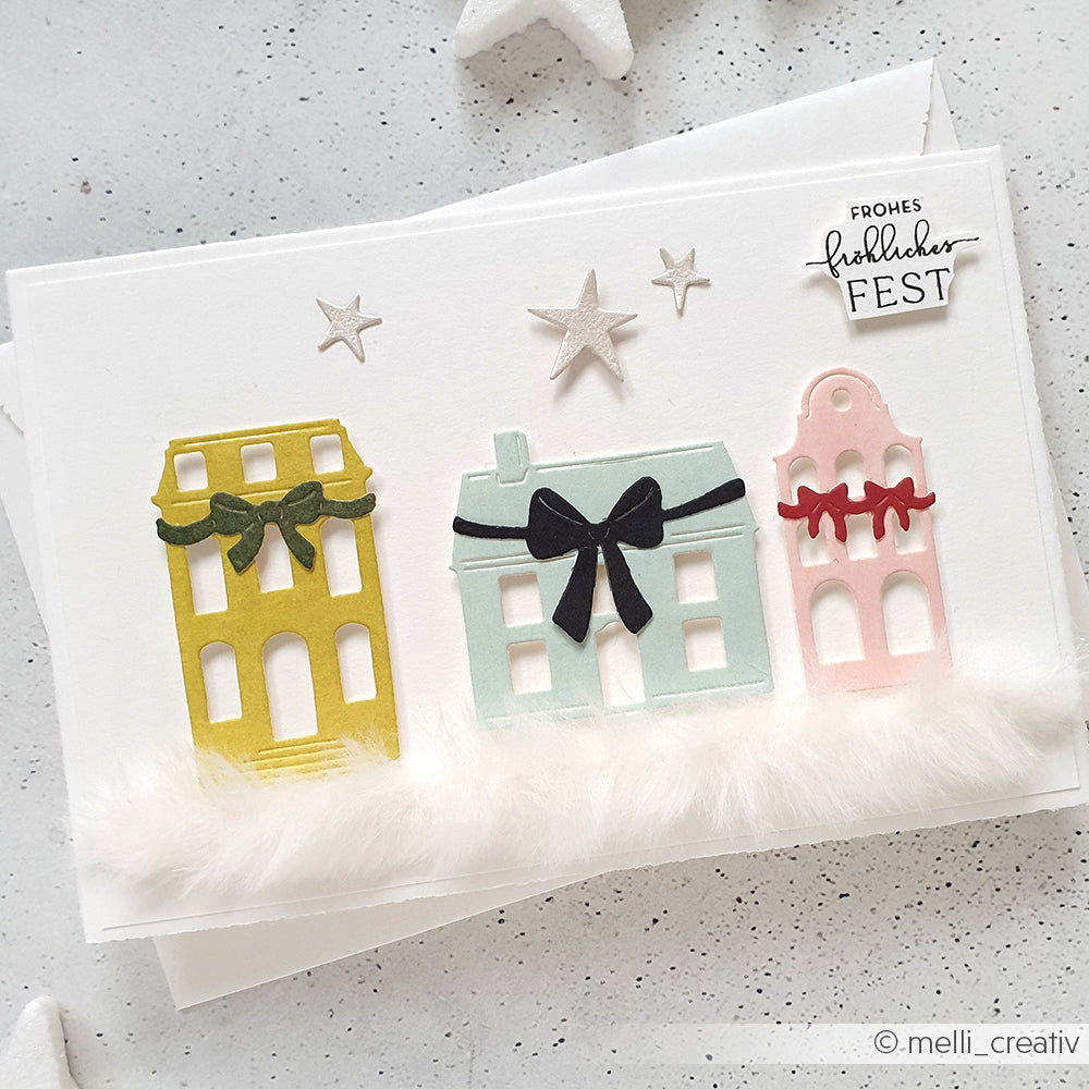 Handgefertigte Weihnachtskarte von Alexandra Renke mit drei pastellfarbenen Häusern, Schleifen, Sternen, flauschiger weißer Bordüre und einem festlichen deutschen Text für die Weihnachtszeit.