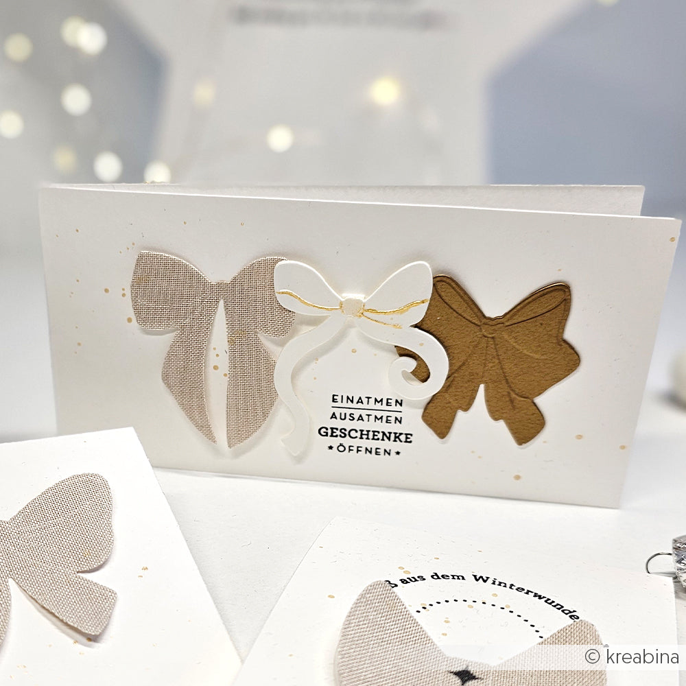 Eine handgefertigte Karte von Alexandra Renke mit drei dekorativen Schleifen, deutschem Text, neutralen Farben, goldenen Akzenten und klaren Photopolymer-Stempeldetails - ideal für die Weihnachtszeit.