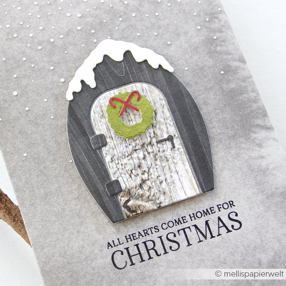 Graue Weihnachtskarte von Alexandra Renke mit einer schneebedeckten gewölbten Tür, grünem Kranz und "ALL HEARTS COME HOME FOR CHRISTMAS". Ideal zum Gestalten mit Ihrem Lieblingsstempel oder Photopolymer-Stempel für ein gemütliches Ambiente.