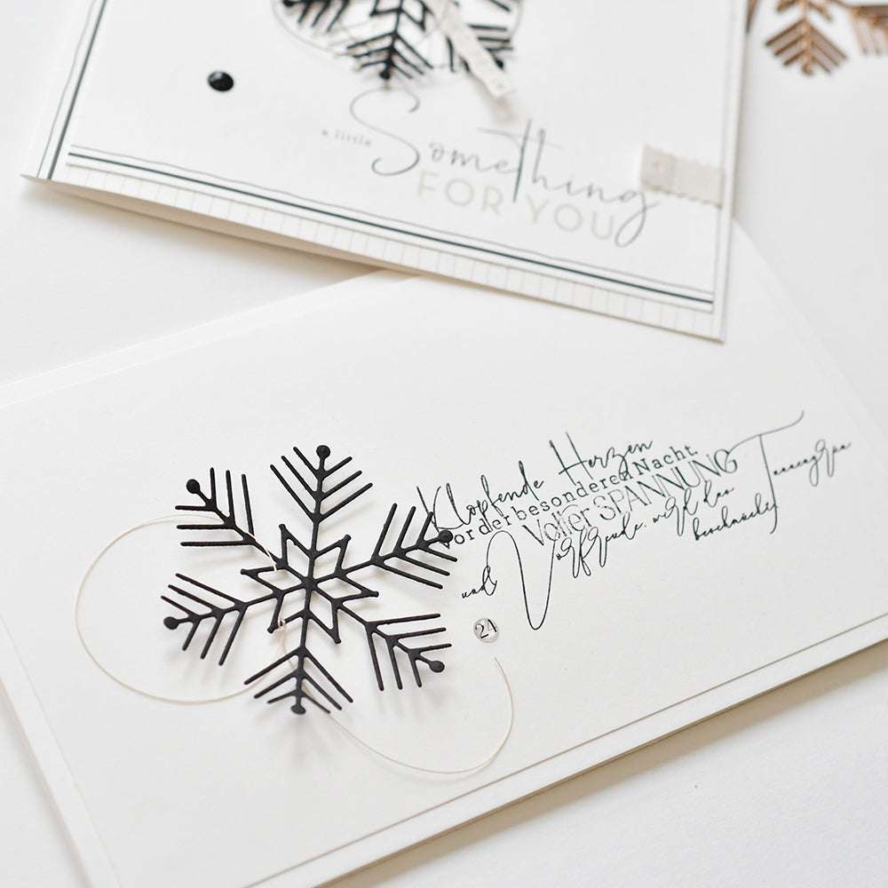 Weiße Karten mit schwarzen Schneeflockenmustern, hergestellt mit Alexandra Renke Motivstanze Eiskristall Stanzen für Papier, zeigen eine elegante kursive Schrift in Englisch und Deutsch.