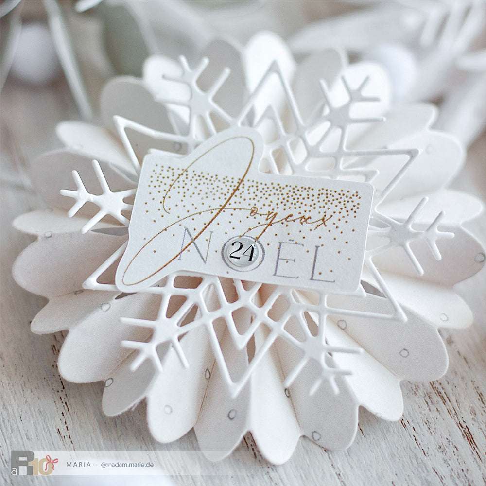 Weiße Schneeflocken aus Papier, hergestellt mit der Motivstanze Eiskristall von Alexandra Renke, mit einem Joyeux Noël Anhänger und der Zahl 24 in der Mitte.