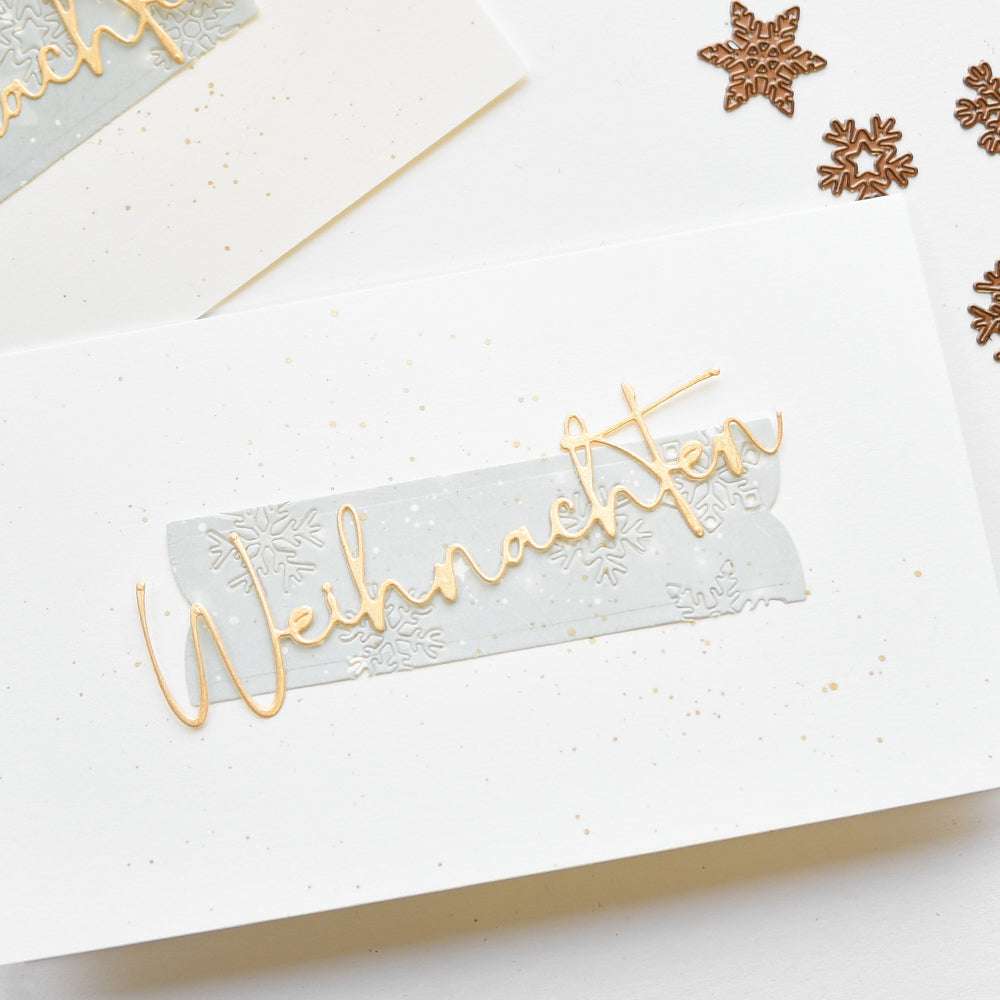 Weiße Karte von Alexandra Renke mit "Weihnachten" in goldener Schrift auf grauem Band, verziert mit Motivstanze Schneeflocken und Schneeflocken-Konfetti - perfekt für festliche Designpapiere.