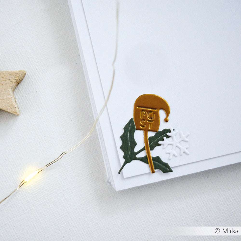 Goldener Briefkasten, grüne Stechpalme und weiße Schneeflockenverzierung auf einem Stapel von Alexandra Renke Designpapieren, akzentuiert mit einer Lichterkette.
