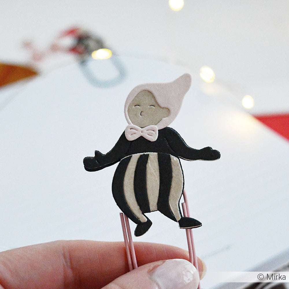 Eine Hand hält einen Clip mit einer niedlichen Cartoon-Figur in schwarz-weißer Kleidung und einer rosa Schleife - hergestellt mit Alexandra Renke Motivstanze Wichtel Snorre für kompatible Stanzsysteme.