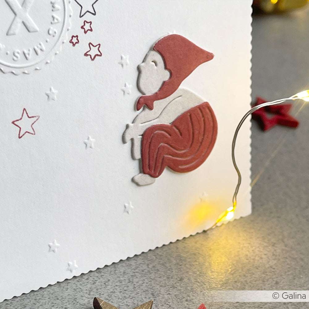 Papierausschnitt einer sitzenden Elfe mit roter Mütze und Hose auf einer weißen Karte, umgeben von Sternen und Lichterketten - gebastelt mit der Motivstanze Wichtel Lars von Alexandra Renke.