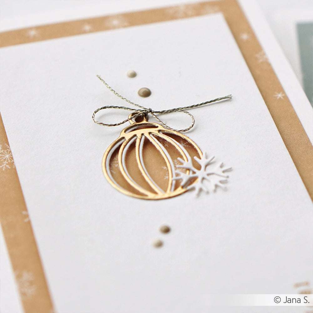 Eine Weihnachtskarte von Alexandra Renke zeigt goldene Ornamente, eine weiße Schneeflocke und eine silberne Schleife, die mit Designpapieren und der Motivstanze Baumkugel gestaltet wurden, auf einem neutralen Hintergrund.