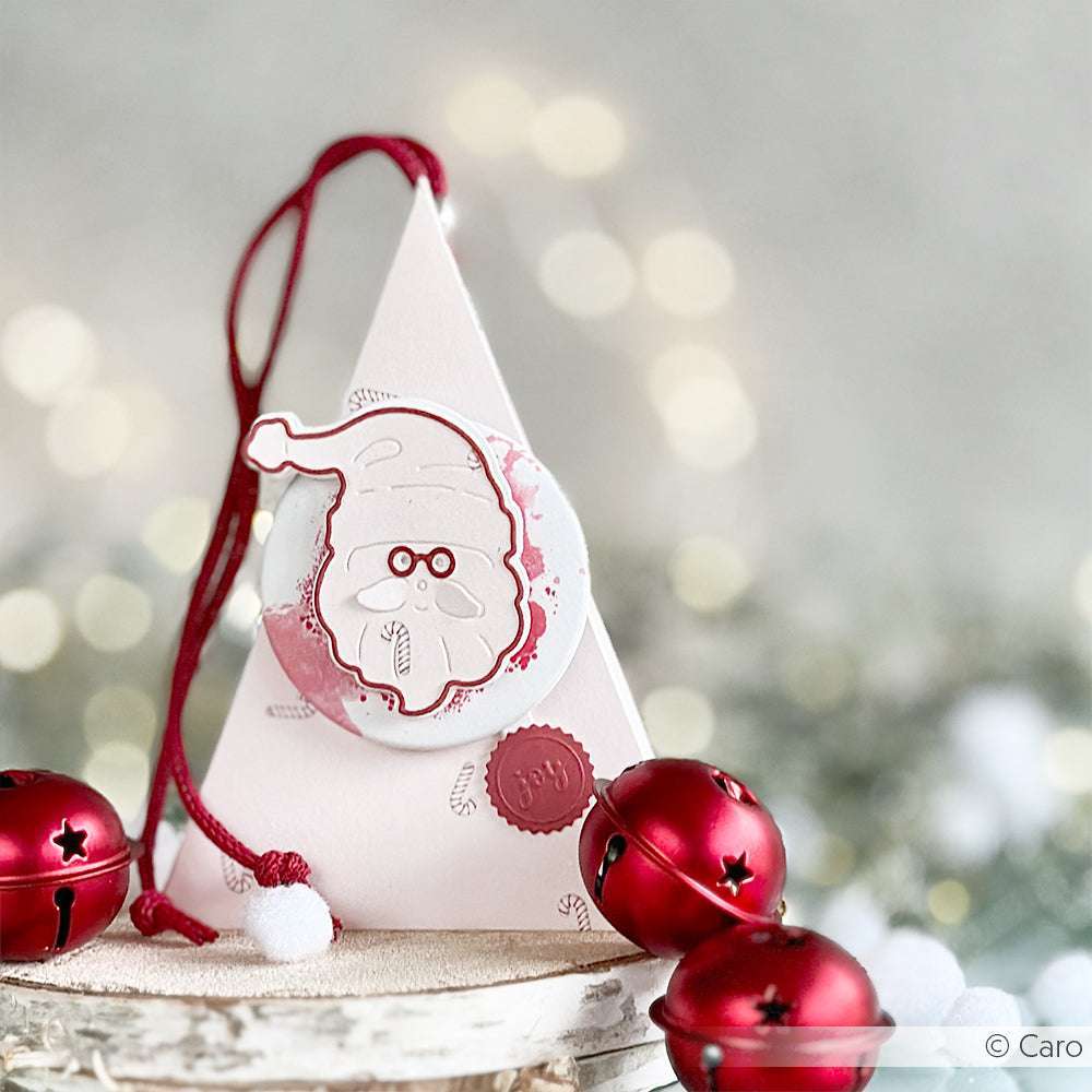 Alexandra Renke Dreieckige Geschenkschachtel mit einem Weihnachtsmann-Anhänger aus Schicht-Weihnachtsmann-Gesicht, verziert mit roter Schnur, Pompon und roten Weihnachtsglocken.