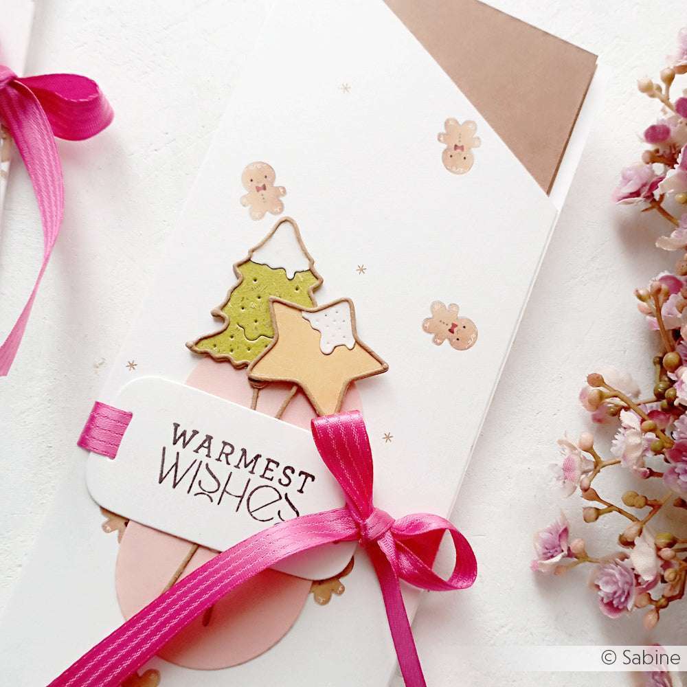 Geschenkkarte von Alexandra Renke mit rosafarbenem Band, Plätzchenbaum und gestanzten Sternen, Text "Warmest Wishes", neben rosa Blumen dargestellt.