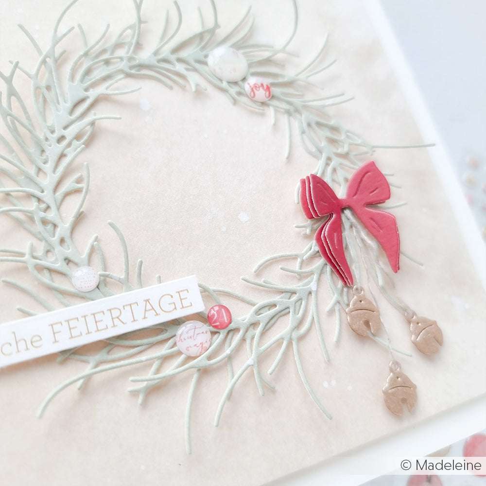 Ein zarter Papierkranz von Alexandra Renke mit roter Schleife, Eichelanhängern und einem "FEIERTAGE"-Anhänger auf hellem Grund - gefertigt mit Motivstanzen und dem Motivstanze Schleifen Quartett für feine Details.