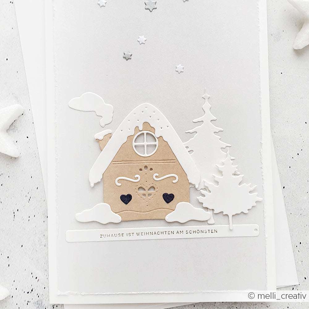 Minimalistische Weihnachtskarte von Alexandra Renke mit einem Lebkuchenhaus aus Kraftpapier, weißen Bäumen, Schnee und verstreuten silbernen Sternen.