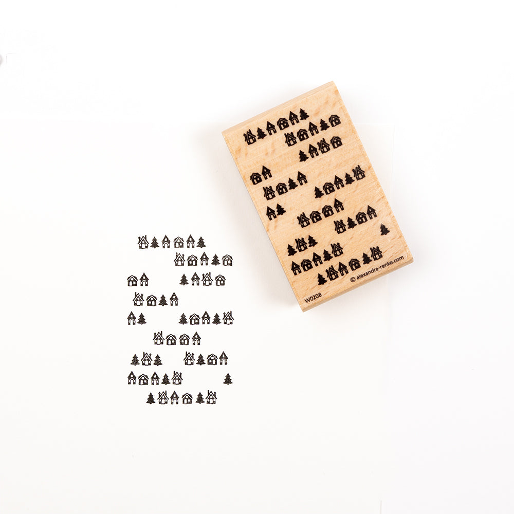 Ein Holzstempel und sein Abdruck mit einem Winterdorf-Motiv, das kleine Häuser und Kiefern zeigt.