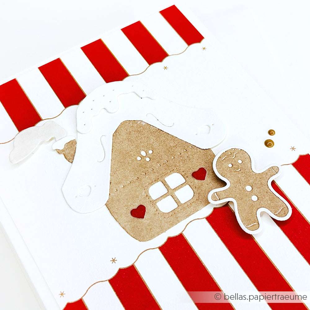 Ein Lebkuchenhaus und eine Person aus Papier, hergestellt mit Alexandra Renke Designpapier und einer Lebkuchenhäuschen-Stanze, vor einem rot-weiß gestreiften Hintergrund.