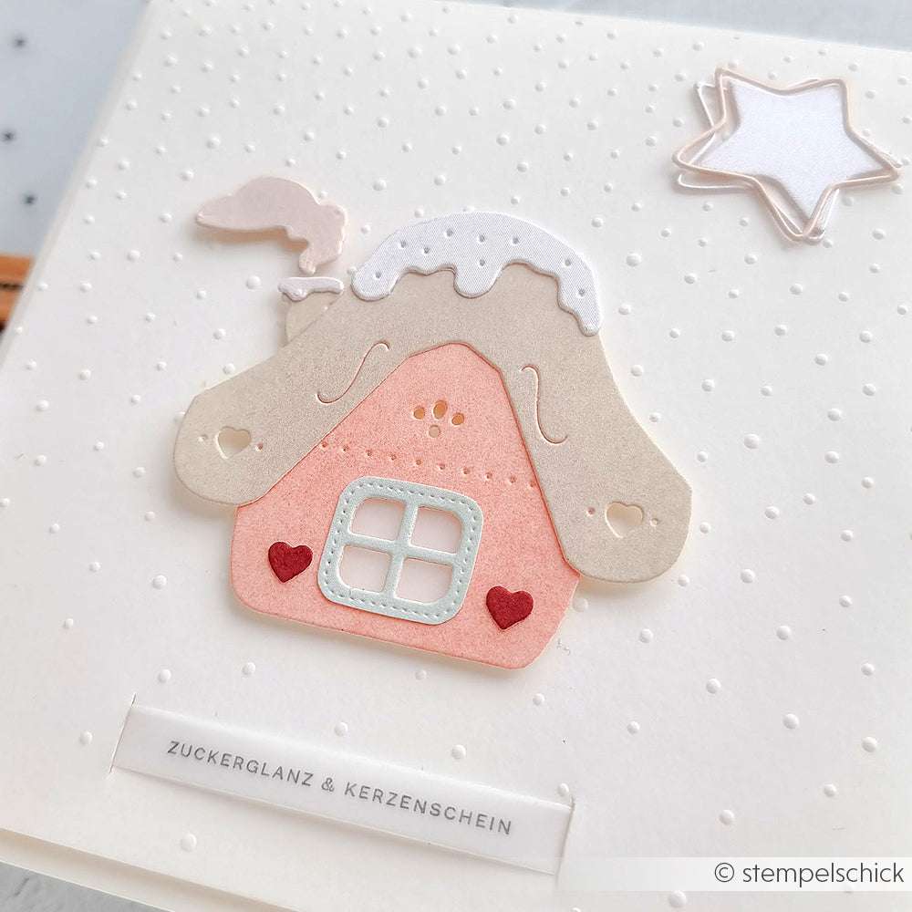 Ein pastellfarbenes Lebkuchenhaus mit Herzen und einem Stern, hergestellt mit der Alexandra Renke Motivstanze Lebkuchenhäuschen, sitzt auf einer weißen Karte mit gepunkteter Struktur.