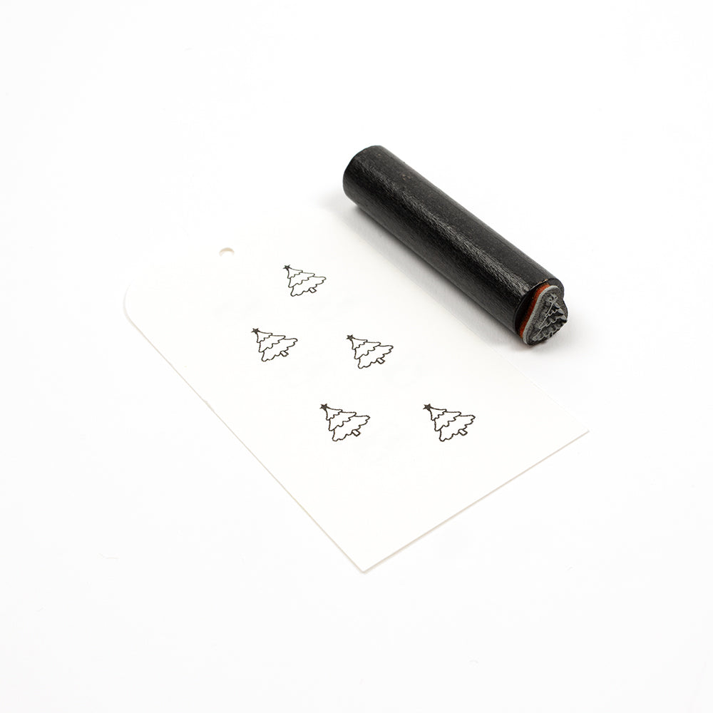 Ein zylindrischer Holzstempel neben einem Papieranhänger, auf dem fünf kleine Weihnachtsbaum-Symbole aufgestempelt sind.