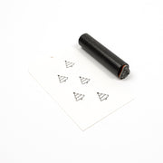 Ein zylindrischer Holzstempel neben einem Papieranhänger, auf dem fünf kleine Weihnachtsbaum-Symbole aufgestempelt sind.
