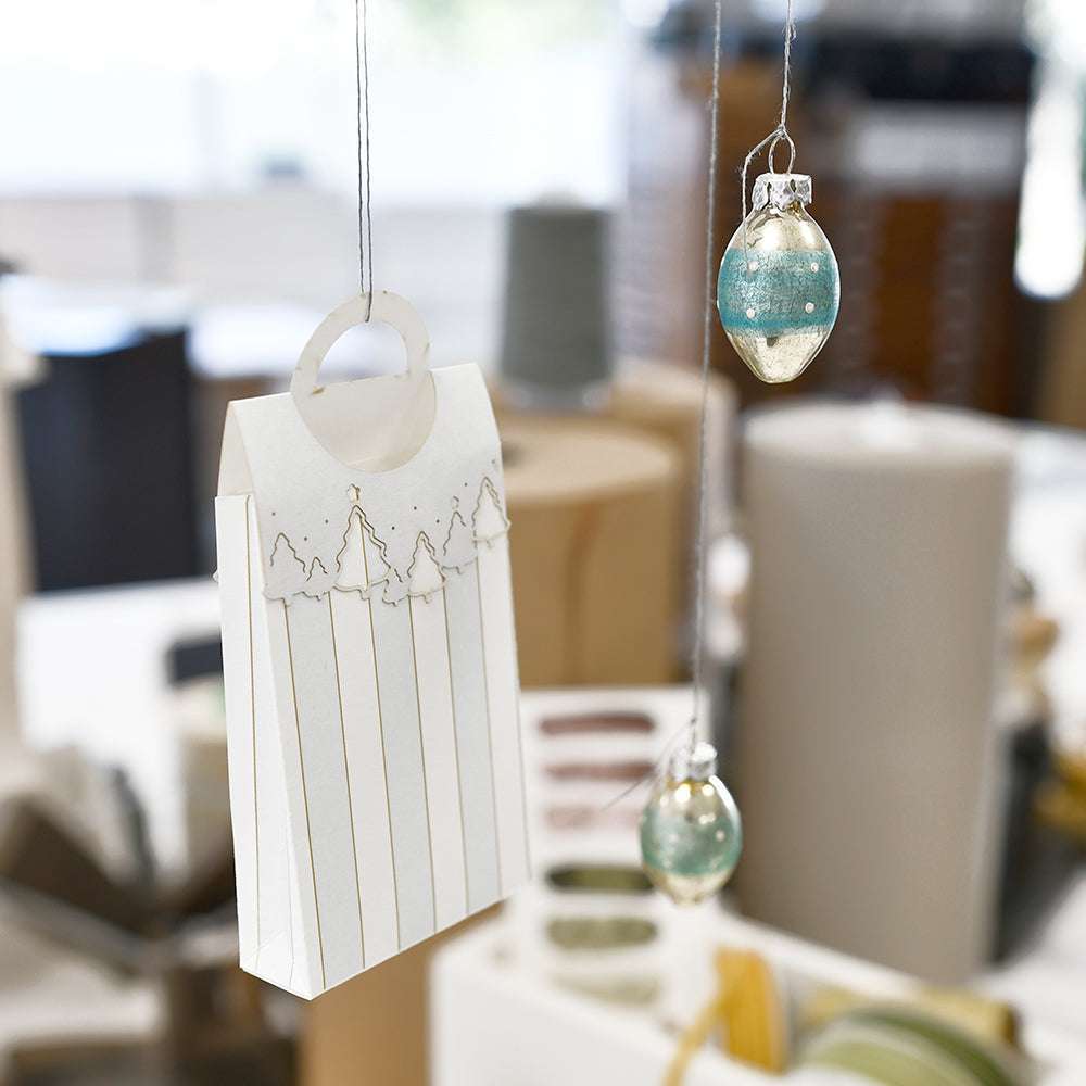 Weiße Geschenktüte von Alexandra Renke mit einem DieCut Systeme-Griff und zwei blauen Hängeornamenten vor einem hellen, leicht verschwommenen Innenhintergrund.