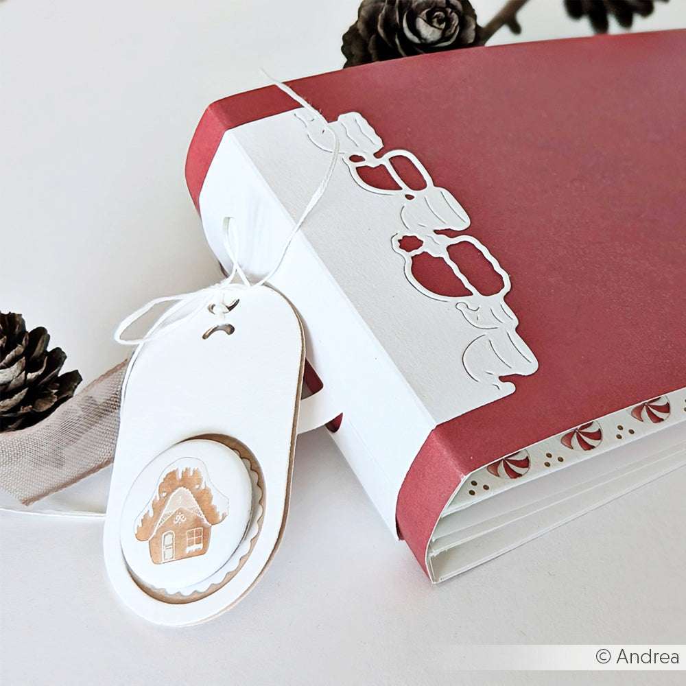 Rot-weißes, handgefertigtes Buch von Alexandra Renke, mit einem Anhänger für ein Winterhäuschen, Tannenzapfen im Hintergrund und festlichen Details, die mit Stanzsystemen und Motivstanze Weihnachtsmützen Dies ausgestanzt wurden.
