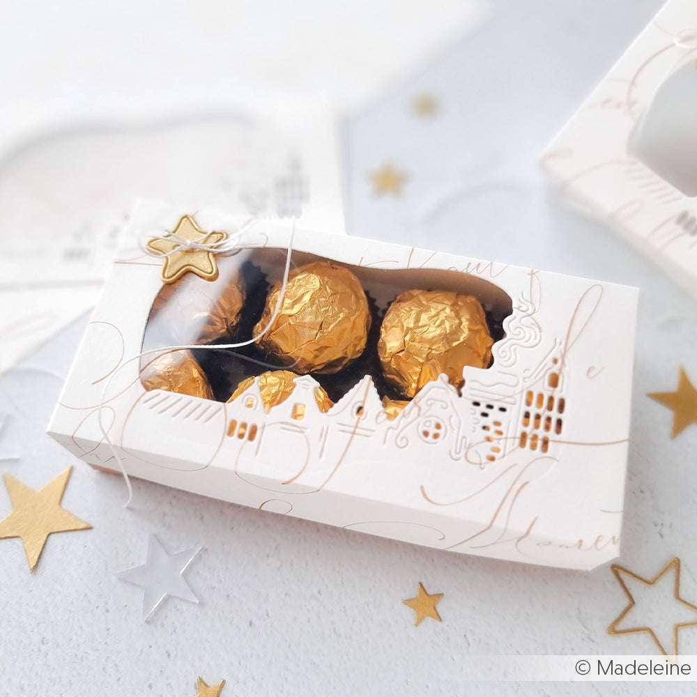 Eine weiße Schachtel mit Sichtfenster zeigt in Gold verpackte Pralinen, umgeben von goldenen und silbernen Papiersternen aus Alexandra Renke Designpapieren und akzentuiert mit Alexandra Renke DieCut Systemen.
