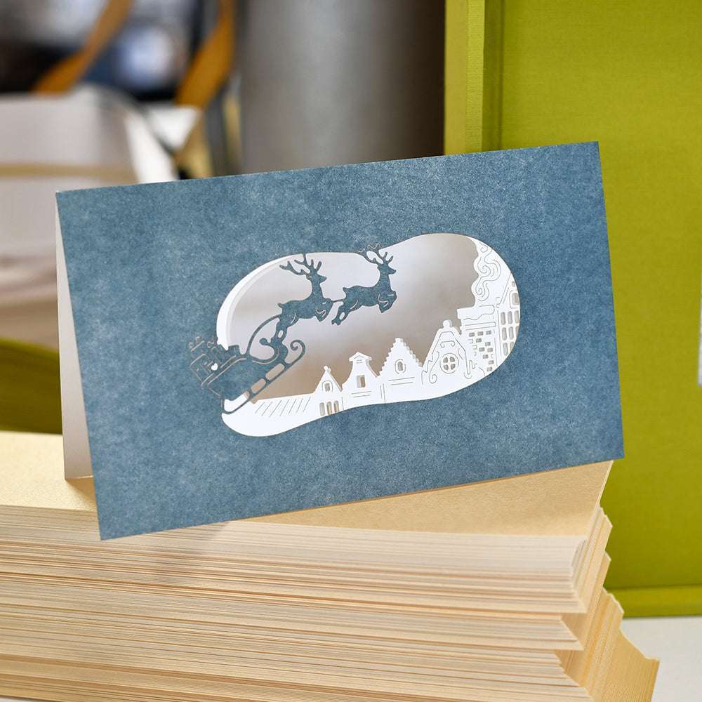 Blaue Weihnachtskarte von Alexandra Renke mit einem Ausschnitt des Weihnachtsmannschlittens und Rentieren, die über Häuser fliegen, dargestellt auf gestapelten Designpapieren.