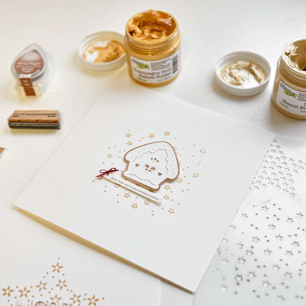 Eine handgefertigte Karte mit einem Lebkuchenhaus, das mit Alexandra Renke Motivstanze Rundes Schicht Lebkuchenhaus gebastelt und mit goldener und weißer Schablonenpaste verziert wurde, ruht schön auf einem weißen Tisch.