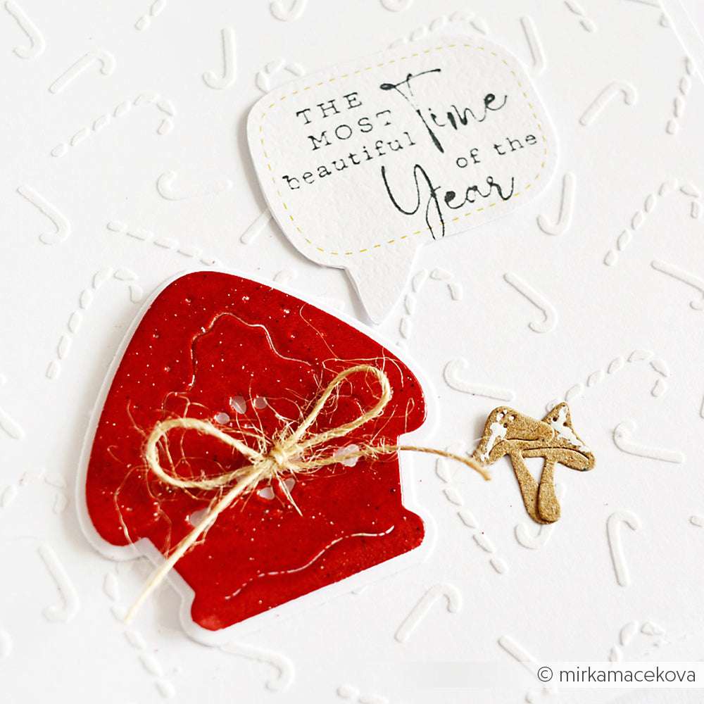 Ein roter Weihnachtsfäustling mit Rankenschleife und goldenem Anhänger, gefertigt mit der Motivstanze Rundes Schicht Lebkuchenhaus von Alexandra Renke, sitzt neben einer Sprechblase mit der Aufschrift "Die schönste Zeit des Jahres".