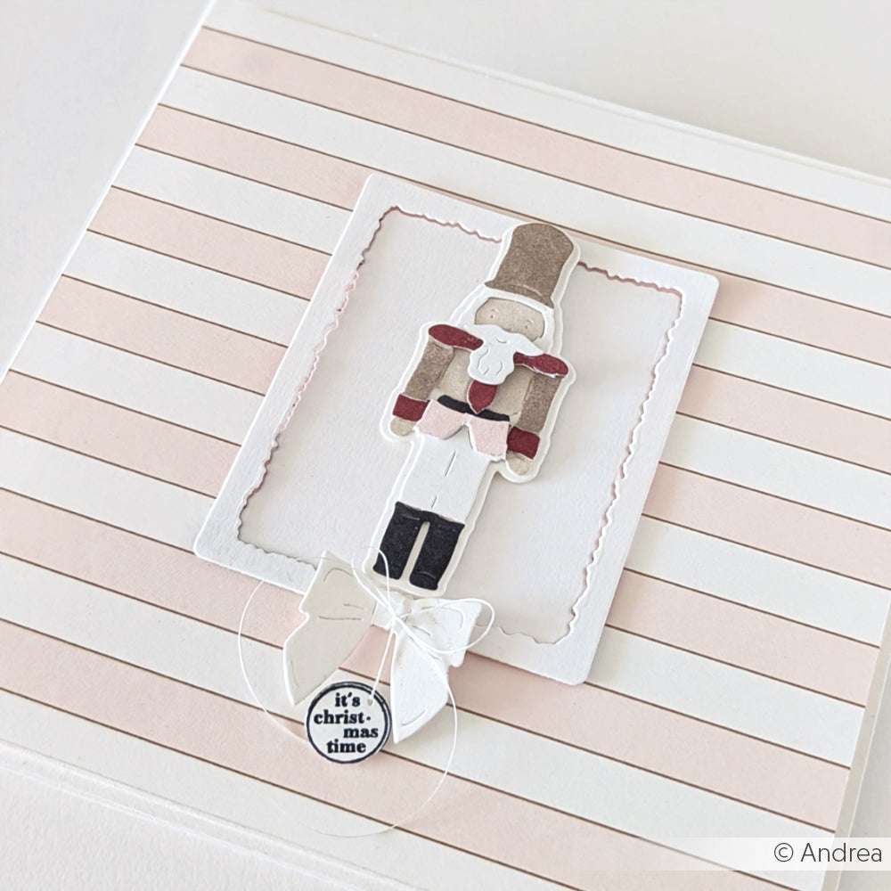 Eine handgefertigte Weihnachtskarte von Alexandra Renke, hergestellt mit Designpapier, zeigt ein Nussknacker-Punsch-Motiv, eine festliche Schleife und "It's Christmas time" auf einem gestreiften Hintergrund.