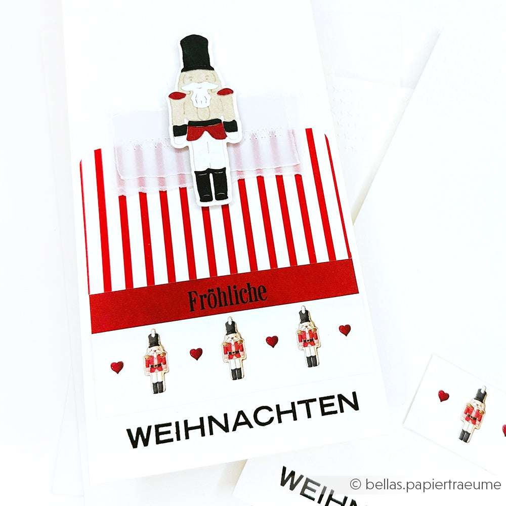 Eine festliche Karte mit einem Nussknacker aus der Alexandra Renke Motivstanze Nussknacker, roten Streifen, kleinen Herzen und dem Text "Fröhliche Weihnachten" auf hochwertigem Designpapier.