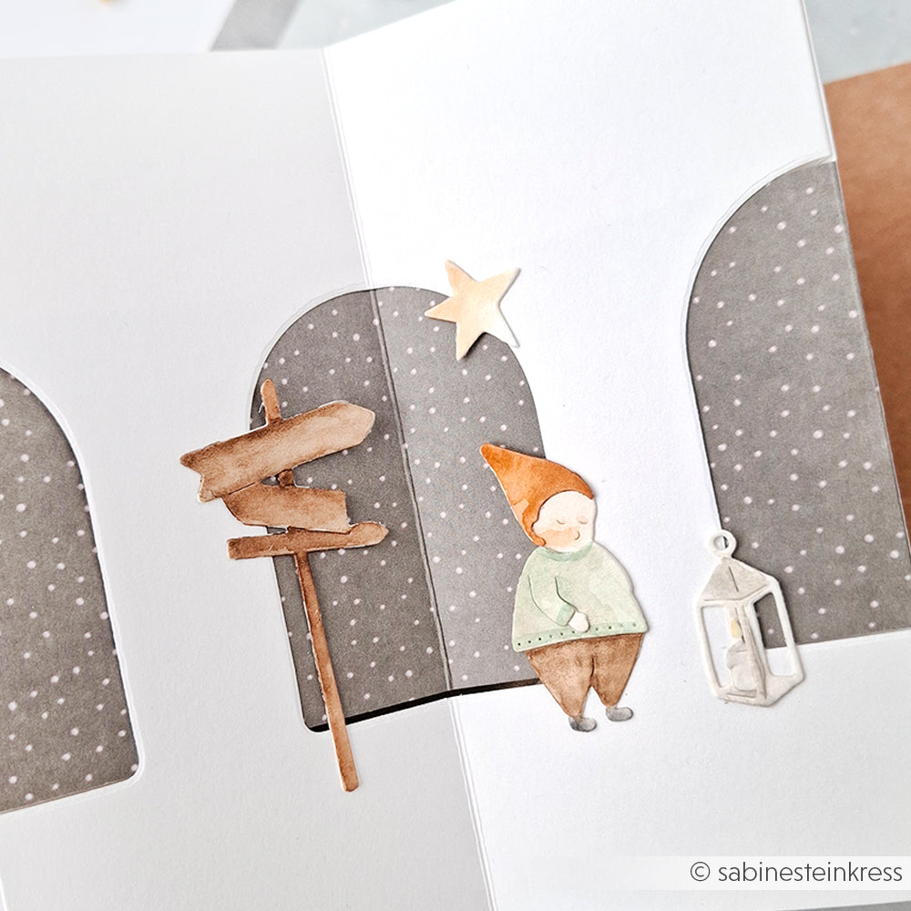 Eine handgefertigte Karte von Alexandra Renke zeigt einen Gnom, einen hölzernen Wegweiser, eine Laterne und einen Stern auf einem gepunkteten grauen Hintergrund.