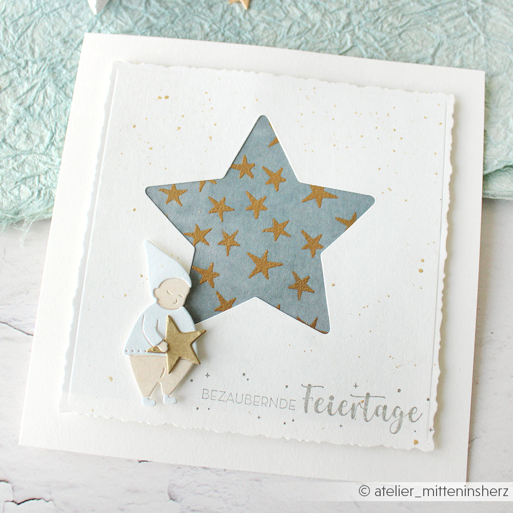Handgeschöpfte Karte mit Alexandra Renke Designpapieren, einem sternförmigen Ausschnitt, goldenen Sternen und einer kleinen Papierfigur aus der Motivstanze Wichtel Sternwanderung, die einen Stern hält.