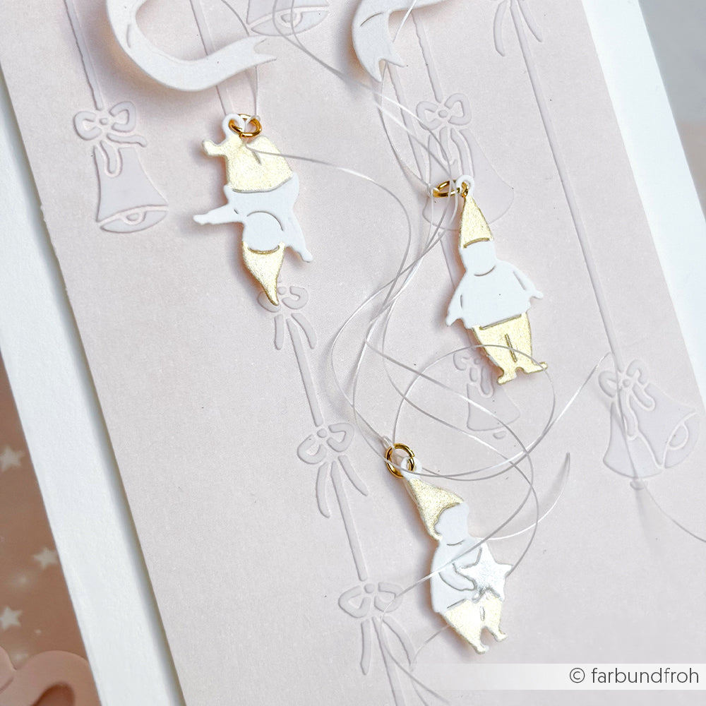 Zwei kleine gold-weiße Wichtelanhänger von Alexandra Renke hängen an klaren Fäden inmitten von Glocken- und Schleifenmotiven - ideal als Motivstanze Kleine Wichtel Anhänger, um Ihren Designpapieren einen skurrilen Charme zu verleihen.