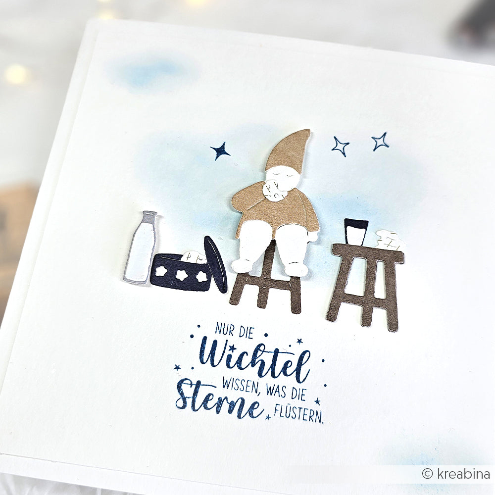 Ein Papierzwerg von Alexandra Renke sitzt auf einem Hocker neben Milch und einem Koffer, mit deutschem Text, Sternen und verspielten Akzenten aus der Motivstanze Wichtel Nascherei Dies Stanzsysteme.