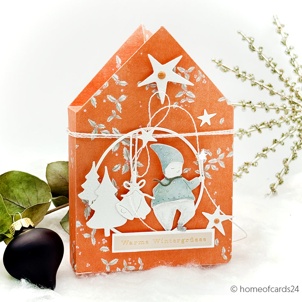 Orangefarbene Karte in Hausform von Alexandra Renke, gestaltet mit DieCut Systemen und Motivstanze Zauber-Wichtel, mit weißen Winterdekor-Sternen, Bäumen und einem Wichtel, der ein Etikett "Wintergrüße" hält.