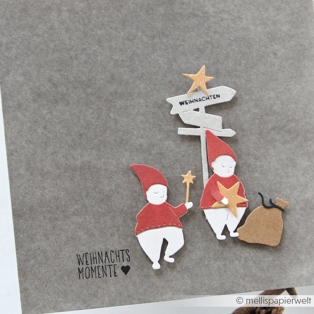 Zwei Papierzwerge mit Weihnachtsmützen, hergestellt mit den Zauber-Wichtel-Stanzen von Alexandra Renke, stehen neben einem Wegweiser und einem Sack auf einer Karte mit der Aufschrift "Weihnachtsmomente".