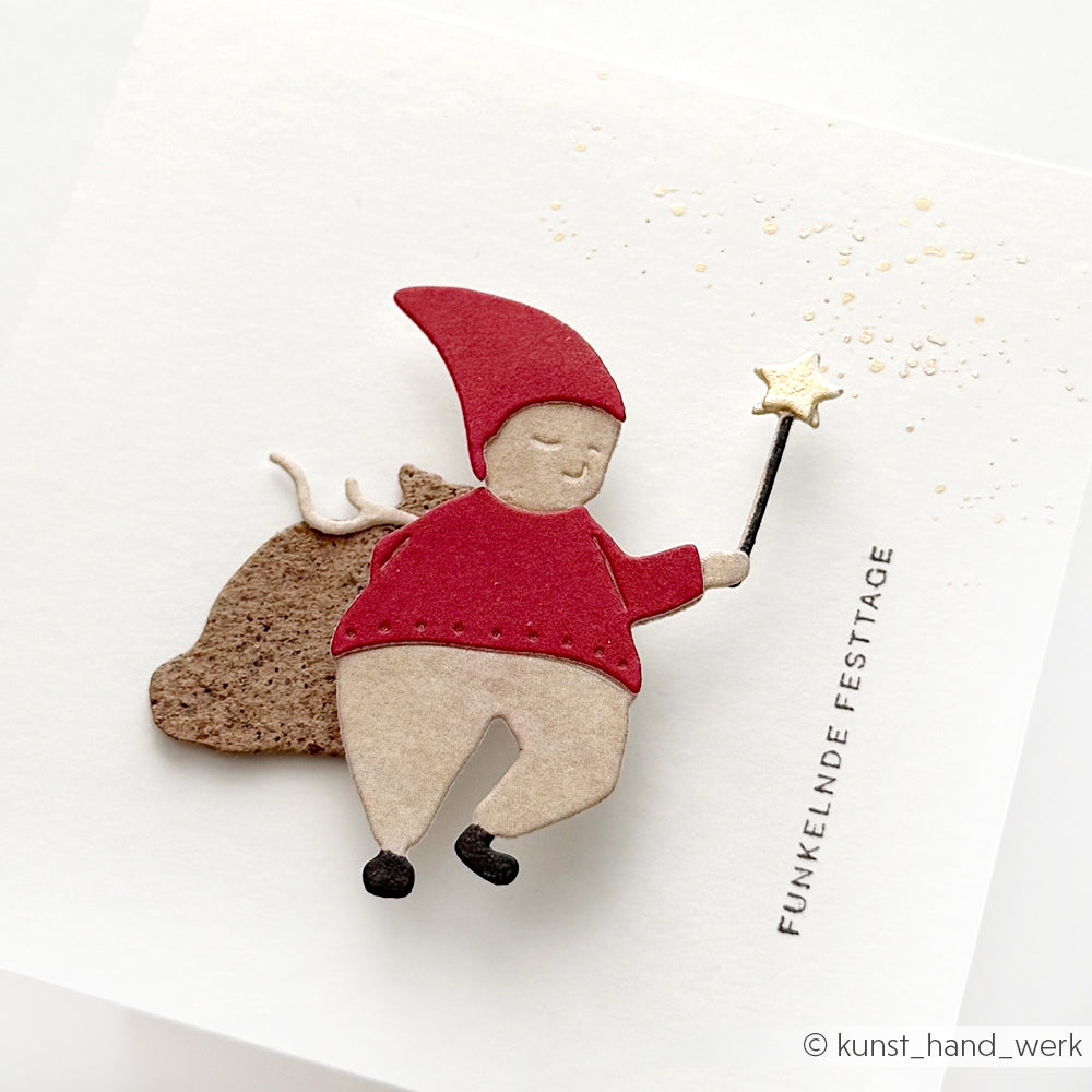 Scherenschnitt eines Zwerges mit Zauberstab und Sack, hergestellt mit Alexandra Renke Designpapiere und Motivstanze Zauber-Wichtel DieCut System, auf einer weißen Karte mit der Aufschrift "Funkelnde Festtage".