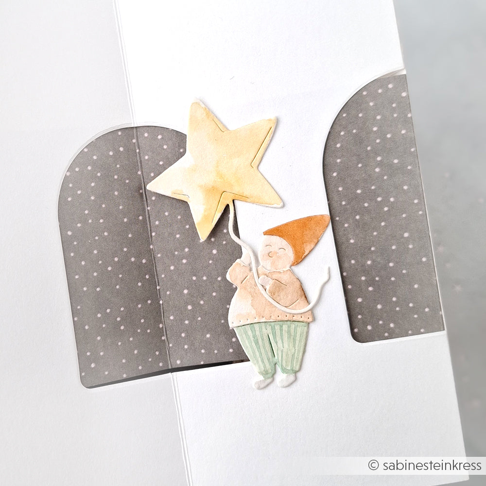 Ein Gnom mit Sternballon, gebastelt mit der Alexandra Renke Motivstanze Wichtel Sternzauber, steht vor einem gepunkteten Bogen auf einer weißen Karte.