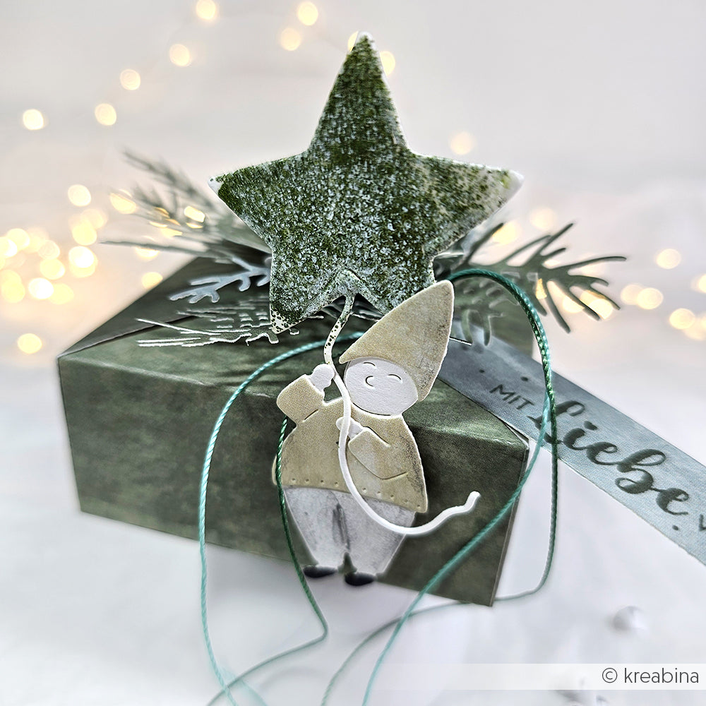 Grüne Geschenkschachtel mit Stern und lächelnder Elfe, hergestellt mit Alexandra Renke Motivstanze Wichtel Sternzauber und festlichen Designpapieren, gebunden mit Schleife, vor einem sanft verschwommenen hellen Hintergrund.