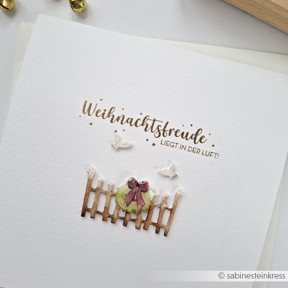 Handgemachte Karte von Alexandra Renke mit "Weihnachtsfreude liegt in der Luft!" über einer Motivstanze Kleiner Zaun mit Kranz, mit zwei weißen Vögeln und festlichen Designpapieren.