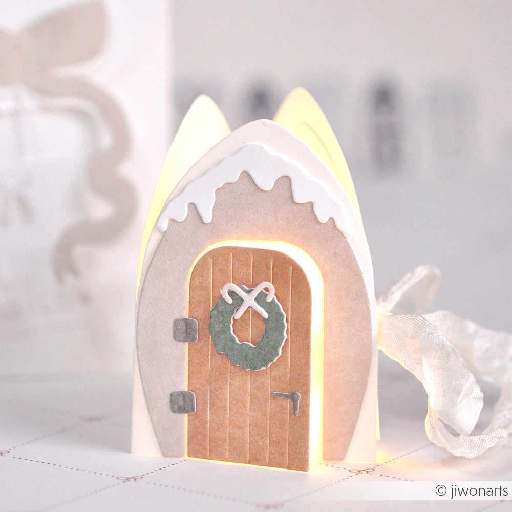 Ein kleines Papierhaus von Alexandra Renke mit einer "Motivstanze Kleine Tür mit Kranz" erstrahlt im Inneren in weichem Licht.