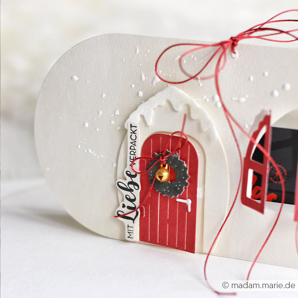 Nahaufnahme einer festlichen weißen Karte mit roter Tür und rotem Kranz, gestaltet mit der Motivstanze Kleine Tür mit Kranz und Designpapieren von Alexandra Renke, verziert mit einer goldenen Glocke und einer roten Schnurschleife.