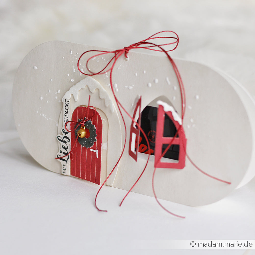 Ovale Weihnachtskarte von Alexandra Renke, gestaltet mit Designpapieren. Mit roter Tür, Kranz, verschneiten Fenstern aus der Motivstanze Kleines Fenster und roter Schleife auf strahlend weißem Hintergrund.