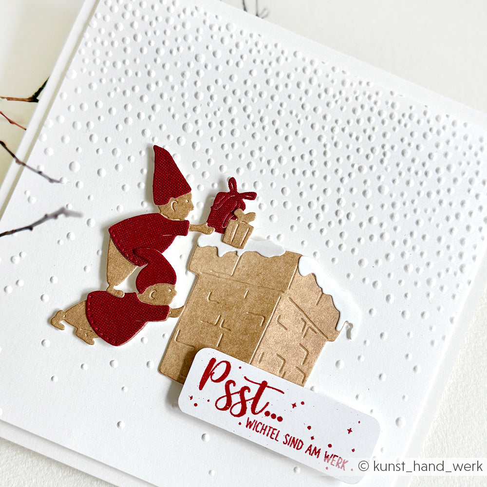 Eine handgemachte Karte mit zwei roten Elfen, einem Haus, einem Geschenk, einer Schneetextur und dem Text "Psst... Wichtel sind am Werk", hergestellt mit Alexandra Renke Designpapieren und der Motivstanze Kleiner Wichtel-Kamin.