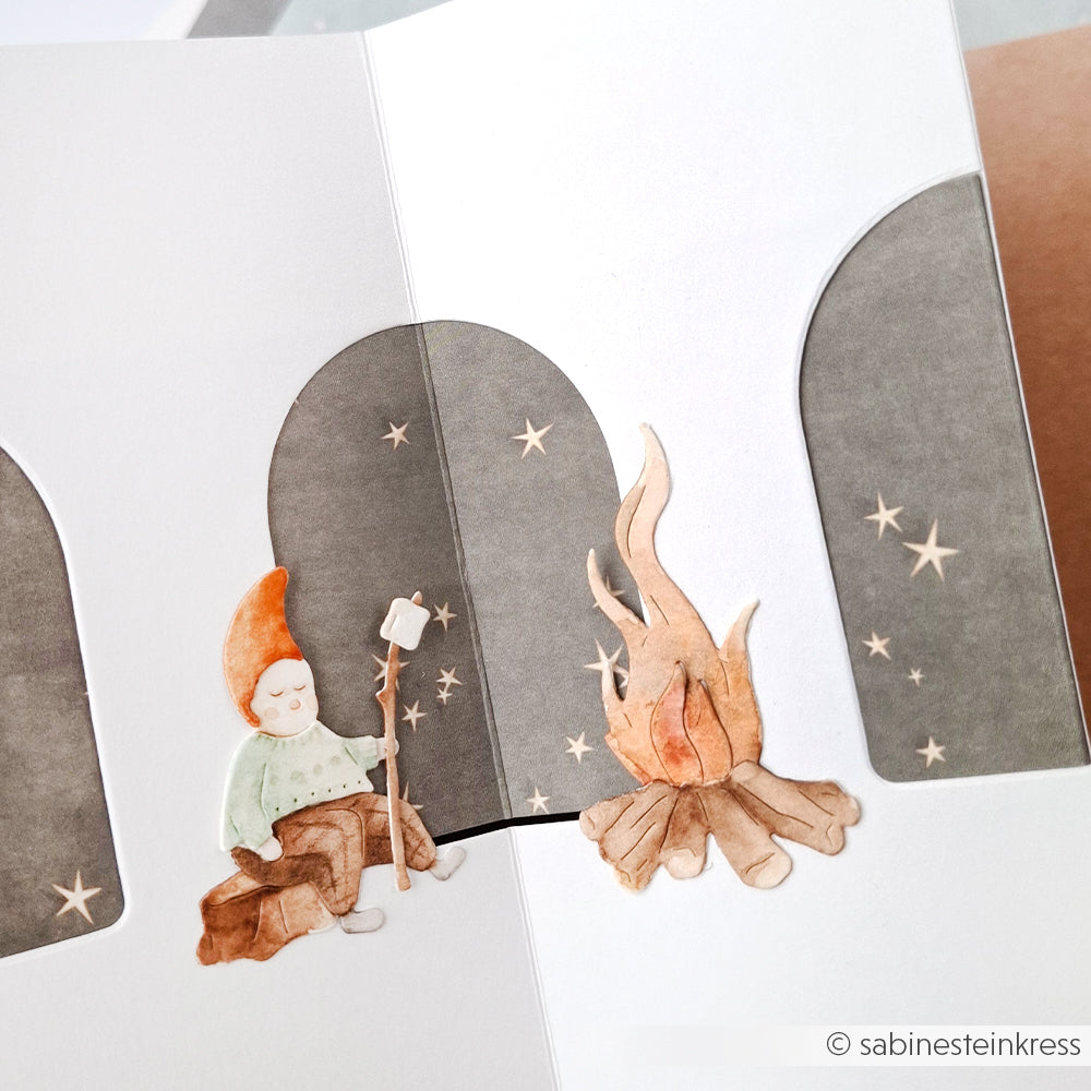 Illustration eines kleinen Wichtels, der mit Alexandra Renke's Designpapieren und der Motivstanze Wichtel am Lagerfeuer ein Marshmallow röstet und dabei die Sterne funkeln lässt.