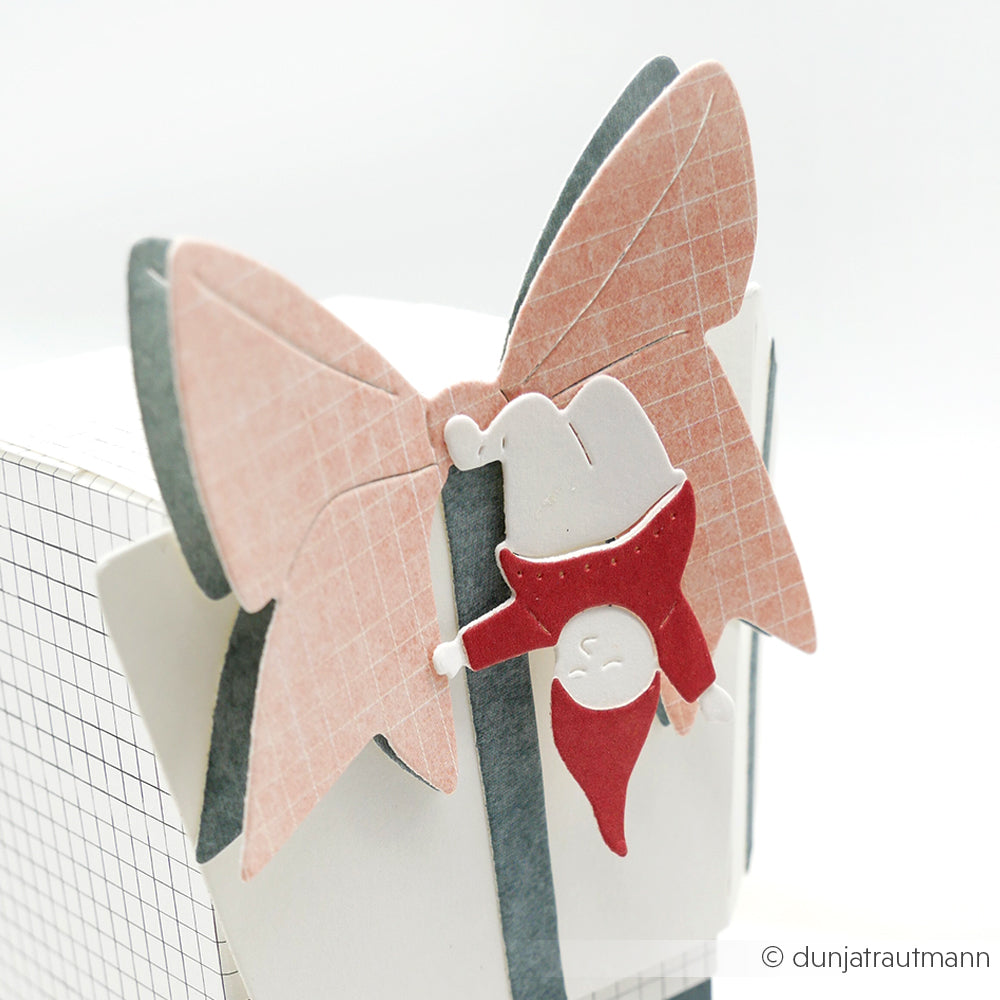 Ein Papierzwerg hängt kopfüber an einer großen rosa-grauen Schleife auf einer karierten Geschenkbox, die mit der Motivstanze Wichtel mit großem Geschenk Dies und Stanzsysteme von Alexandra Renke gebastelt wurde.