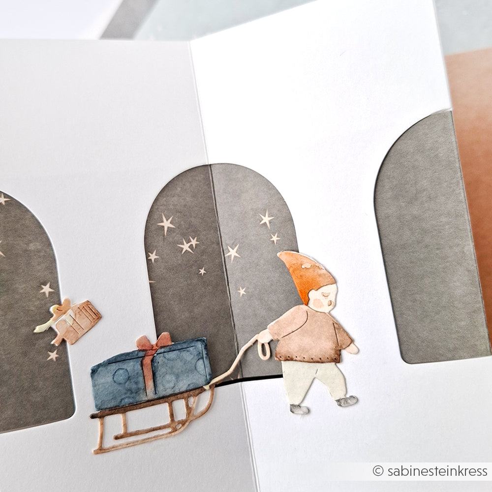 Handgemachte Karte mit einem Alexandra Renke Motivstanze Wichtel Geschenke-Lieferdienst Ausschnitt eines Kindes, das ein Geschenk auf einem Schlitten zieht, mit funkelnden Sternen im Hintergrund.